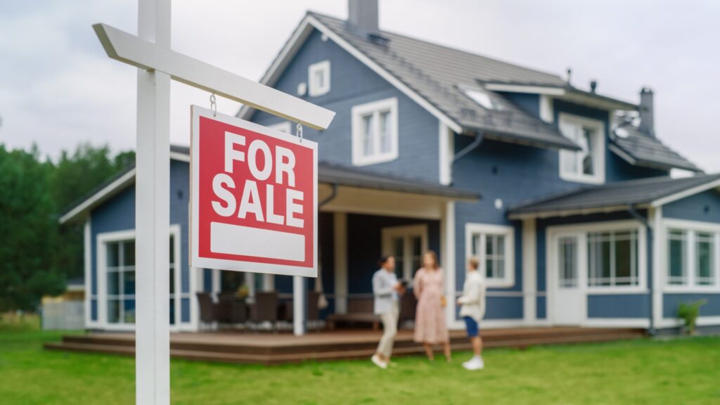 A For Sale sign stands in the yard of a blue house while three people discuss the sale in the background.