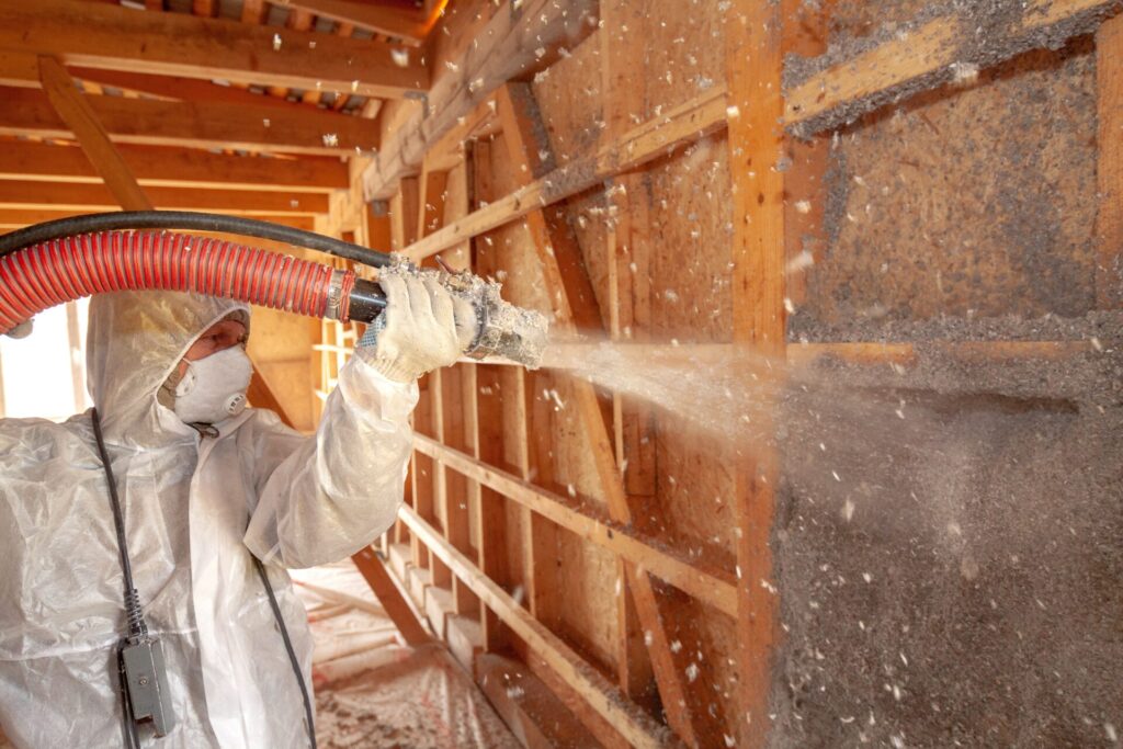 A worker wearing protective gear spraying cellulose insulation onto an unfinished wall 
