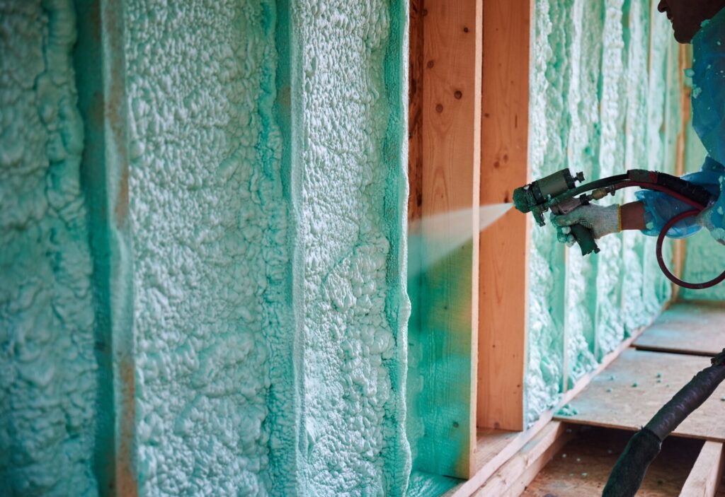 A worker wearing protective gear uses a spray gun to install green spray foam insulation 