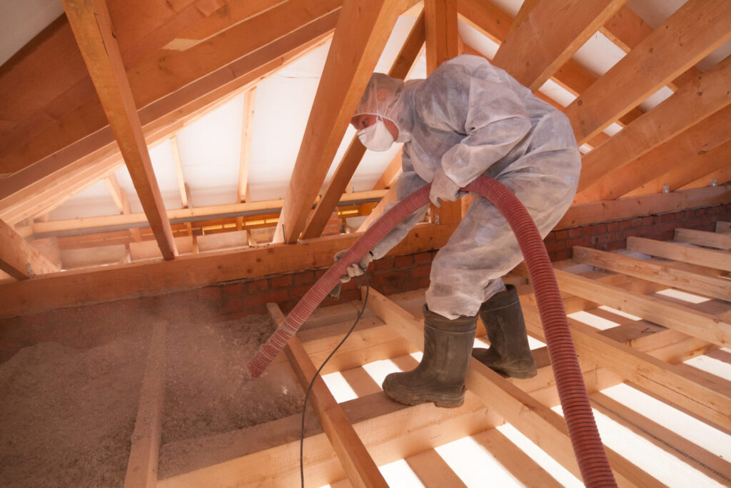 A worker wearing personal protective equipment uses a specialized hose to spray cellulose insulation into an unfinished attic