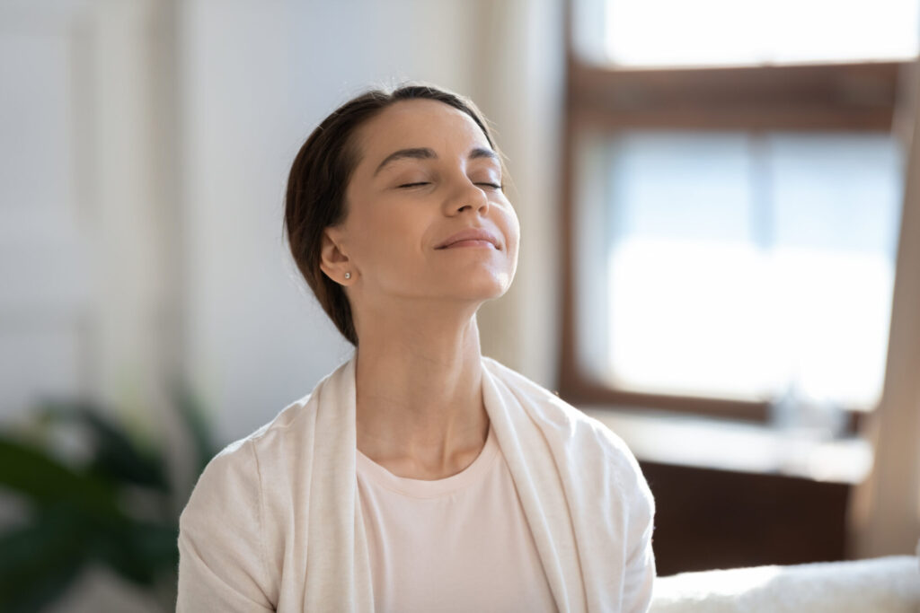 A young woman takes a deep breath indoors, in front of a window.
