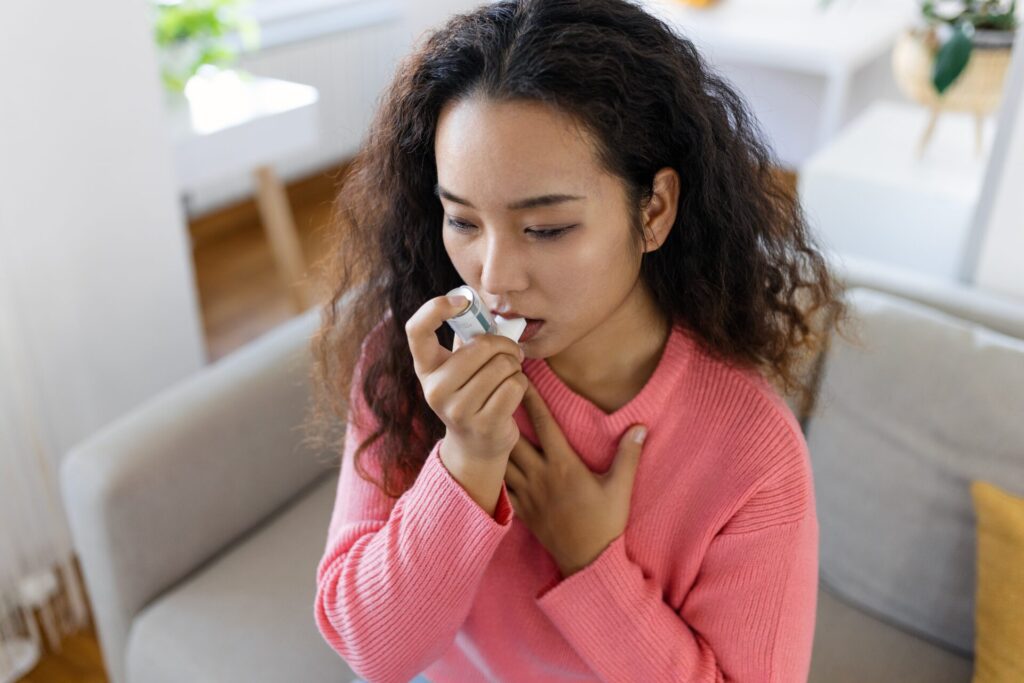 A woman in a pink sweater sits on a gray couch with an asthma inhaler in her mouth  