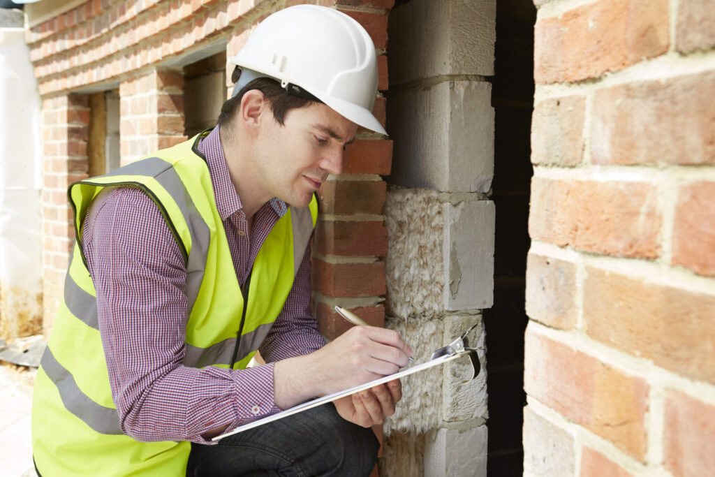 A man in a hardhat and yellow vest writes on a clipboard next to a brick wall