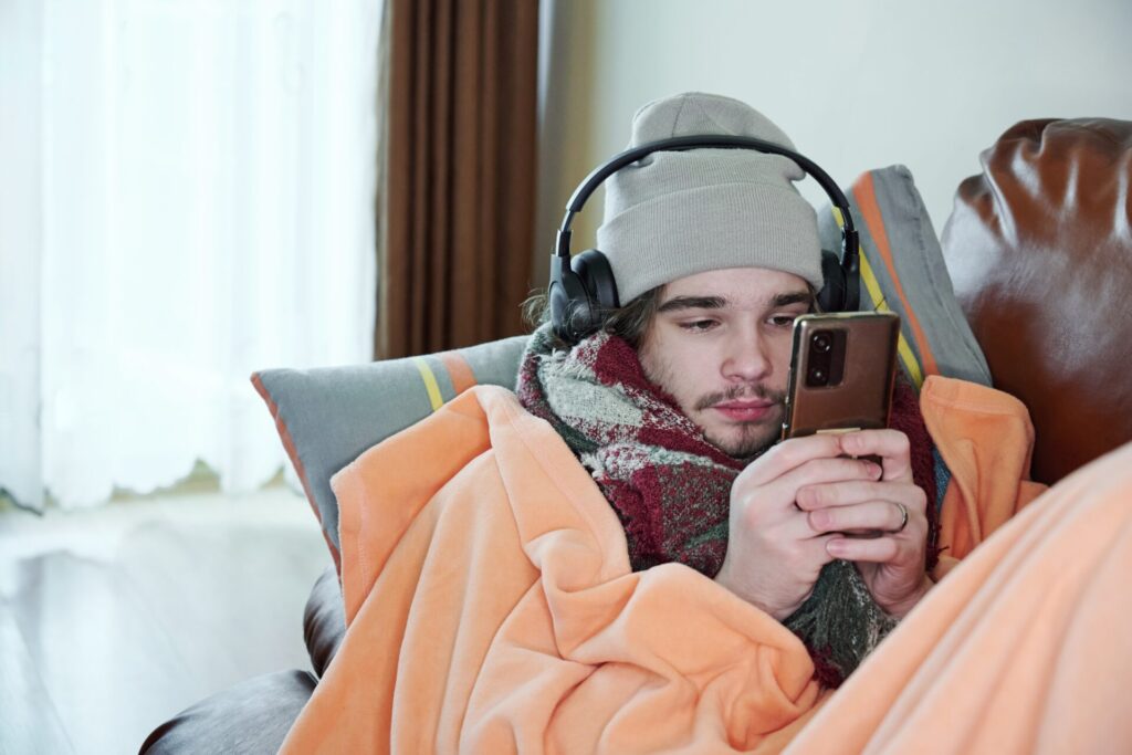 A young man looks at his smartphone on a couch while wearing a beanie, headphones, and wrapped in a scarf and blankets.