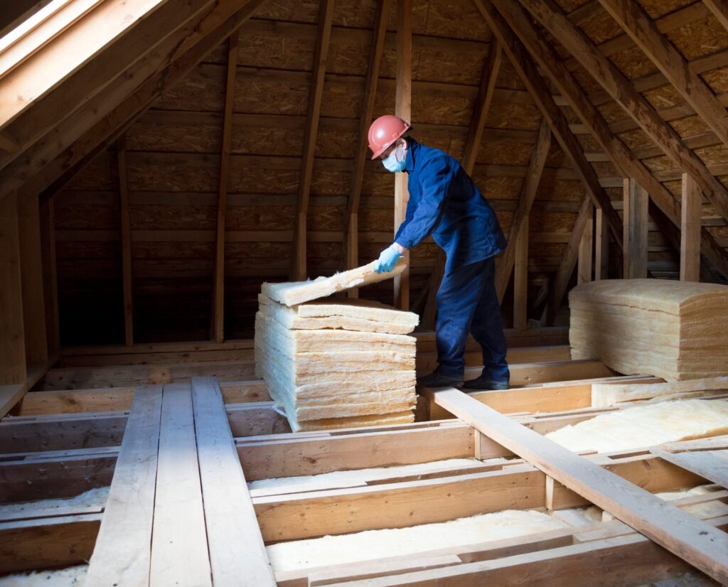 A worker in a blue jumpsuit and protective gear installs insulation in an attic