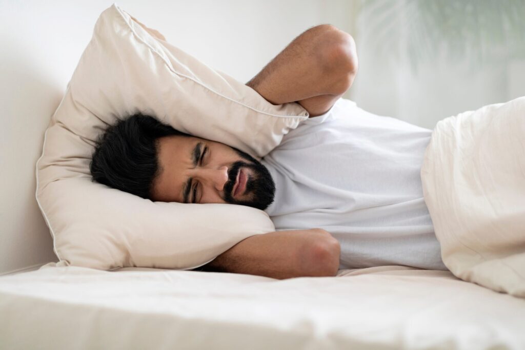 A man with dark hair and a beard lies in bed and covers his ears with a pillow in a brightly lit room