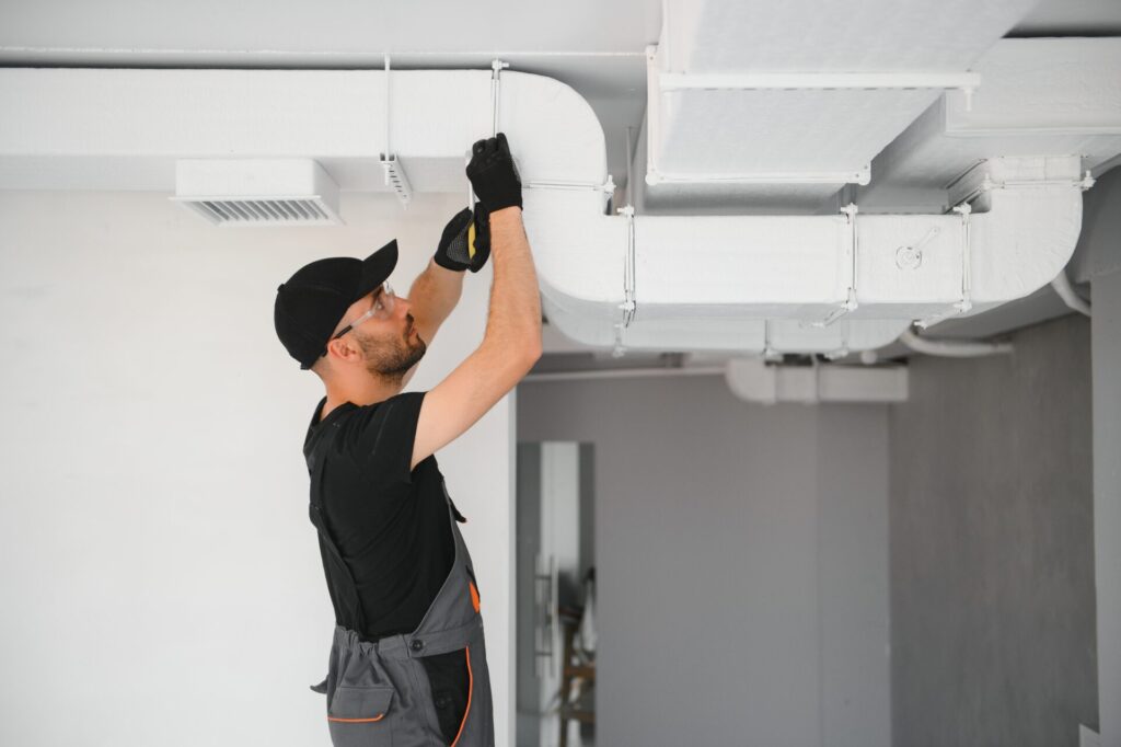 A worker in coveralls works on a white air duct overhead
