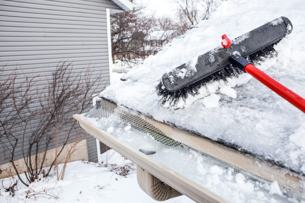 A broom with a red handle sweeps snow off a roof while the gutters are blocked with ice
