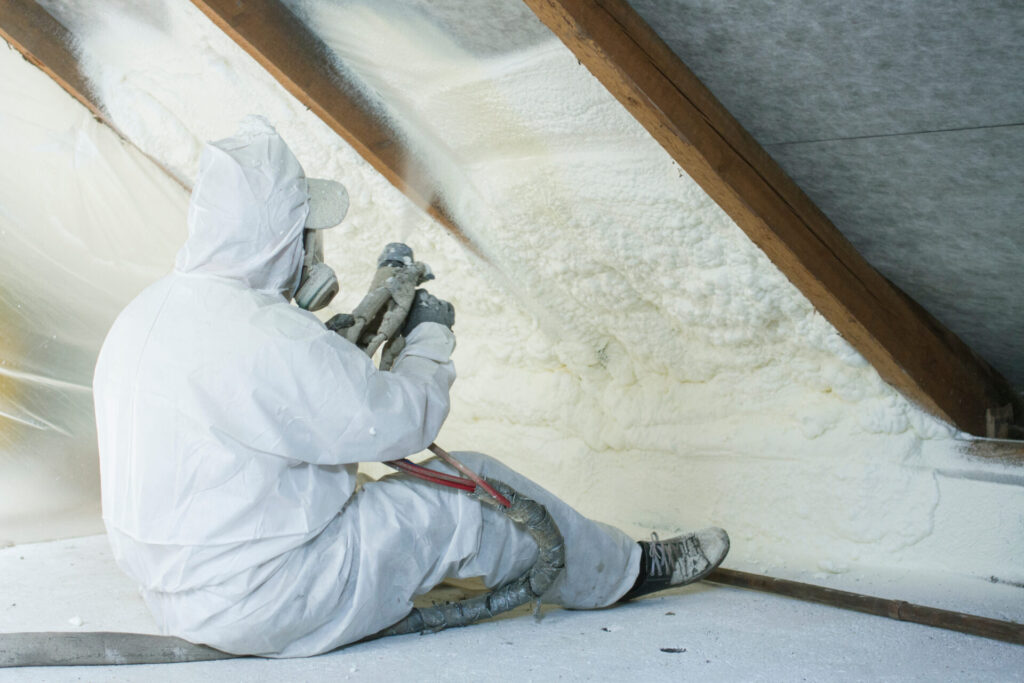 A technician wearing white protective gear and a respirator mask applies spray foam insulation in an attic space