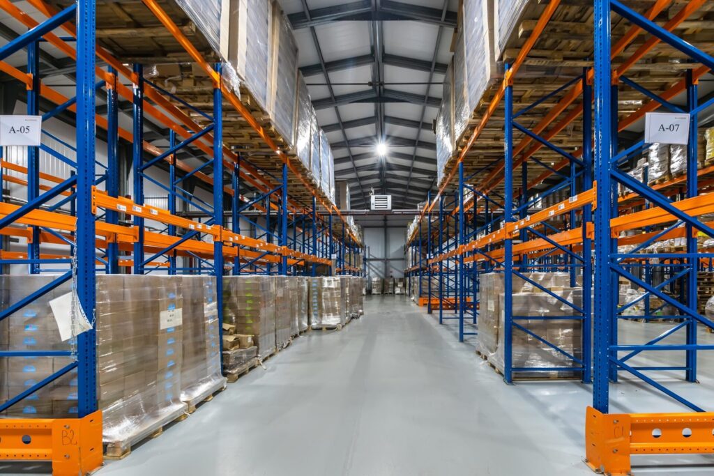 The aisle of a brightly lit commercial warehouse is lined with blue and orange shelves stacked with cardboard boxes