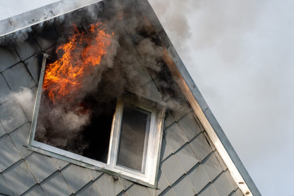  Flames and smoke come out of the attic window of a gray house