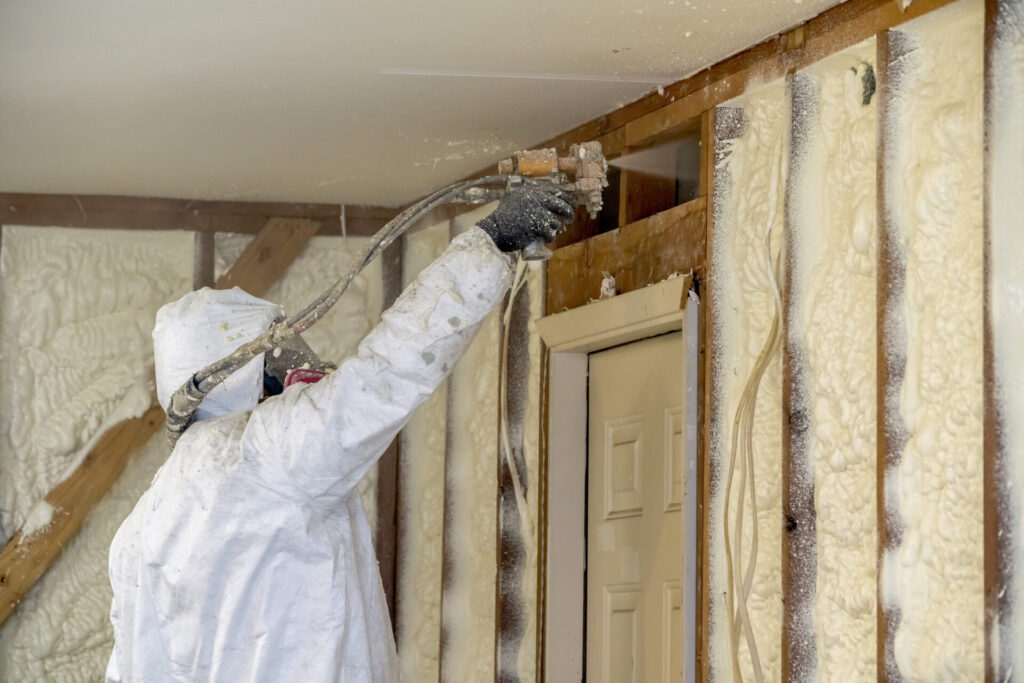 A person wearing personal protective equipment, including coveralls and a respirator sprays insulation foam into a wall frame