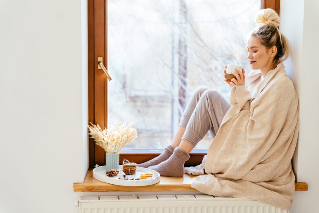 A blonde woman wrapped in a blanket sits near a window with wintery weather outside