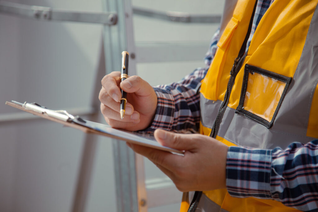 A man wearing a plaid shirt and orange safety vest holds a pen and clipboard
